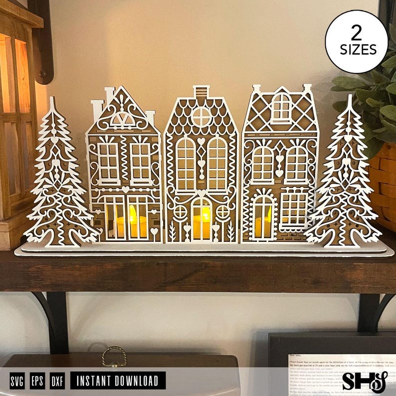 May include: A laser-cut wooden gingerbread village display featuring three houses and two pine trees with intricate white filigree detailing. The structures are arranged on a flat base, with yellow LED tea lights glowing inside the houses.