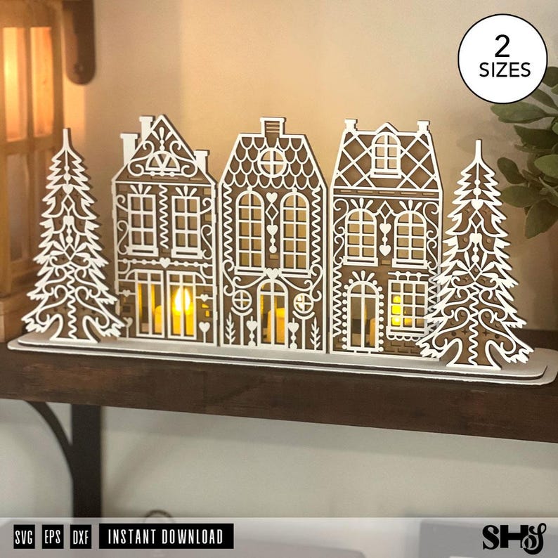 May include: A laser-cut wooden decorative display featuring three gingerbread-style houses flanked by two pine trees. The white-painted intricate lattice patterns overlay the brown wood. A circular badge in the corner reads 2 SIZES.