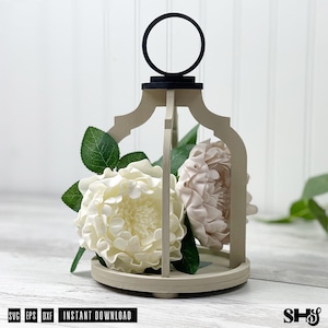 May include: A beige wooden lantern frame with a black circular handle at the top, containing two large white and pale pink artificial flowers with green leaves. The text SVG EPS DXF INSTANT DOWNLOAD is printed at the bottom left corner.
