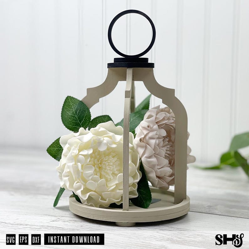 May include: A beige wooden lantern frame with a black circular handle at the top, containing two large white and pale pink artificial flowers with green leaves. The text SVG EPS DXF INSTANT DOWNLOAD is printed at the bottom left corner.