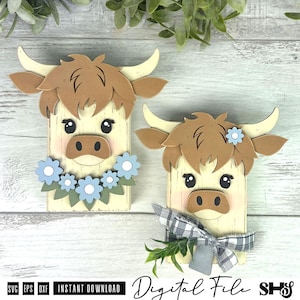 Pu&ograve; includere: Due sagome in legno di mucche highland con volti color crema, capelli marroni e corna. Una mucca presenta una ghirlanda di fiori blu, mentre l'altra indossa un fiocco a quadri bianco e nero con una foglia verde e un singolo fiore blu.