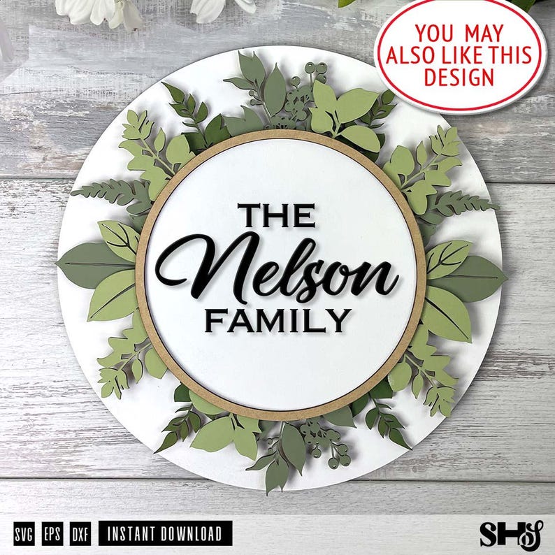 May include: A white round wooden sign with a green leafy wreath around the edge. The wreath is made of layered paper cutouts. The sign has a brown circle in the center with the text "THE Nelson FAMILY" in black.