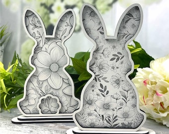 May include: Two wooden cutout bunny shapes with a floral design. The bunnies are standing on a white base. The design is black and white.