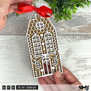 Puede incluir: Adorno de casa de jengibre de madera con intrincados detalles recortados. El adorno presenta una base marr&oacute;n con detalles blancos, incluyendo ventanas, corazones y patrones decorativos. Una cinta roja est&aacute; atada en la parte superior para colgar. El texto "INSTANT DOWNLOAD" es visible.