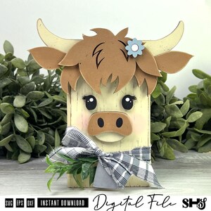 Pu&ograve; includere: Un blocco decorativo in legno a forma di mucca Highland con venature color crema, capelli marroni a strati, corna ricurve e un fiore azzurro. Presenta occhi neri, un muso marrone e un fiocco in tessuto a quadri bianco e nero.