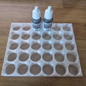 Puede incluir: Un organizador de pintura de plástico blanco con 36 compartimentos redondos. Dos pequeños frascos de pintura acrílica negra están en la fila superior del organizador. Los frascos están etiquetados como "Model Color Acrylic" y tienen el nombre de la marca "Vallejo" en ellos.