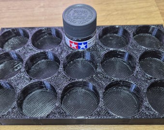 May include: A black 3D-printed paint bottle holder with 15 circular compartments. A bottle of Tamiya paint sits in the center. The paint bottle has a black cap and a label with red and blue stars.