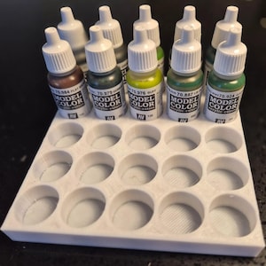 Peut inclure: Un porte-bouteilles de peinture en plastique blanc avec 18 fentes rondes. Le porte-bouteilles est rempli de 8 petites bouteilles de peinture acrylique. Les bouteilles sont étiquetées "Model Color" et ont différentes couleurs, notamment le brun, le vert, le jaune et le bleu.