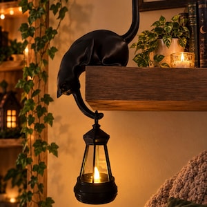 May include: A black cat figurine perched on the edge of a wooden shelf, with its tail extending downwards to hold a black lantern. The lantern features a clear glass pane and contains a warm, glowing LED light that illuminates the surrounding area.