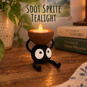 May include: A black, round soot sprite figurine with large white eyes, holding a brown bowl containing a glowing white LED tealight candle. The text SOOT SPRITE TEALIGHT is displayed above the figurine in a dark, simple, sans-serif font.