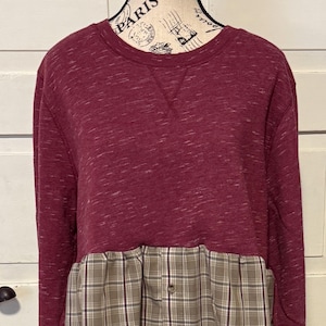 May include: A burgundy sweatshirt with a plaid shirt bottom. The top portion of the sweatshirt is a solid burgundy color with a crew neck. The bottom is a plaid pattern in shades of brown, tan, and gray, with a button closure.
