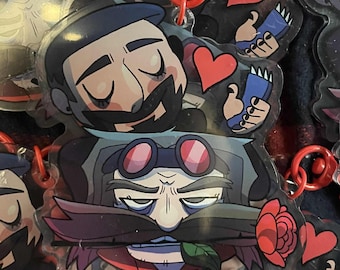 May include: A collection of colorful cartoon character keychains featuring a variety of facial expressions and accessories. The keychains are made of clear acrylic and have a red heart charm attached.