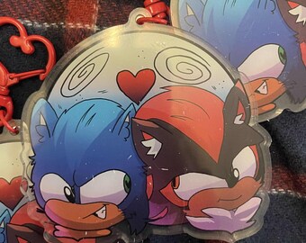 May include: Two cartoon characters, Sonic the Hedgehog and Shadow the Hedgehog, are featured on a clear, round keychain. The characters are drawn in a chibi style and are shown with hearts above their heads. The keychain has a red heart-shaped clip.