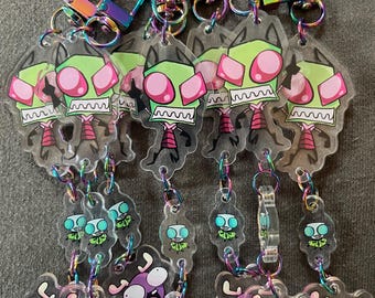 May include: A collection of clear acrylic keychains featuring cartoon characters. The keychains have iridescent clasps and chains. The characters include a green alien with pink eyes, a small blue alien, and a purple creature with a large mouth.