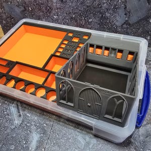 May include: A clear plastic storage container with an orange and black organizer insert. A gray miniature building with arched windows and doors sits inside the container. The organizer has labeled compartments.