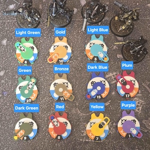 Op de afbeelding: Een verzameling van twaalf ronde plastic speltrackers met draaibare wijzerplaten, elk voorzien van een unieke kleurgecodeerde arm en een genummerd vlak. De trackers zijn gelabeld met kleurnamen zoals Light Green, Gold, Light Blue, Green, Bronze, Dark Blue, Plum, Dark Green, Red, Yellow en Purple, en worden getoond naast miniatuurfiguurtjes.