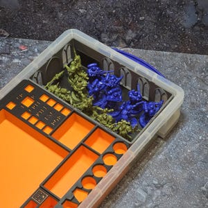 May include: A clear plastic storage container holding miniature figurines in green and blue, alongside a black and orange organizer tray with compartments labeled CP, CRIT, KILL, TAC, and POP, designed for organizing gaming components.
