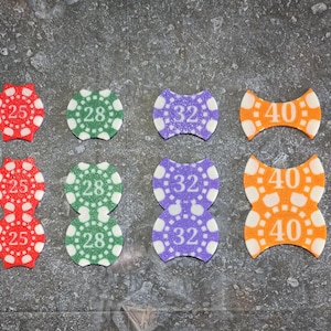 Puede incluir: Una colección de fichas de póquer coloridas con diferentes denominaciones. Las fichas son rojas, verdes, moradas y naranjas, cada una mostrando un número: 25, 28, 32 y 40. Las fichas tienen una superficie texturizada y una forma única.