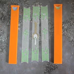 Könnte beinhalten: Draufsicht auf drei transparente, rechteckige Teile mit grünen schaltungsartigen Mustern und einem zentralen goldfarbenen Element. Zwei leuchtend orangefarbene, rechteckige Teile flankieren die transparenten Teile. Es handelt sich um Komponenten oder Zubehör.