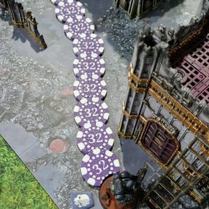May include: A line of purple and white poker chips with the number 32 printed on each chip. The chips are arranged in a line on a gray surface. A miniature figure of a soldier is standing on a brown base next to the chips.
