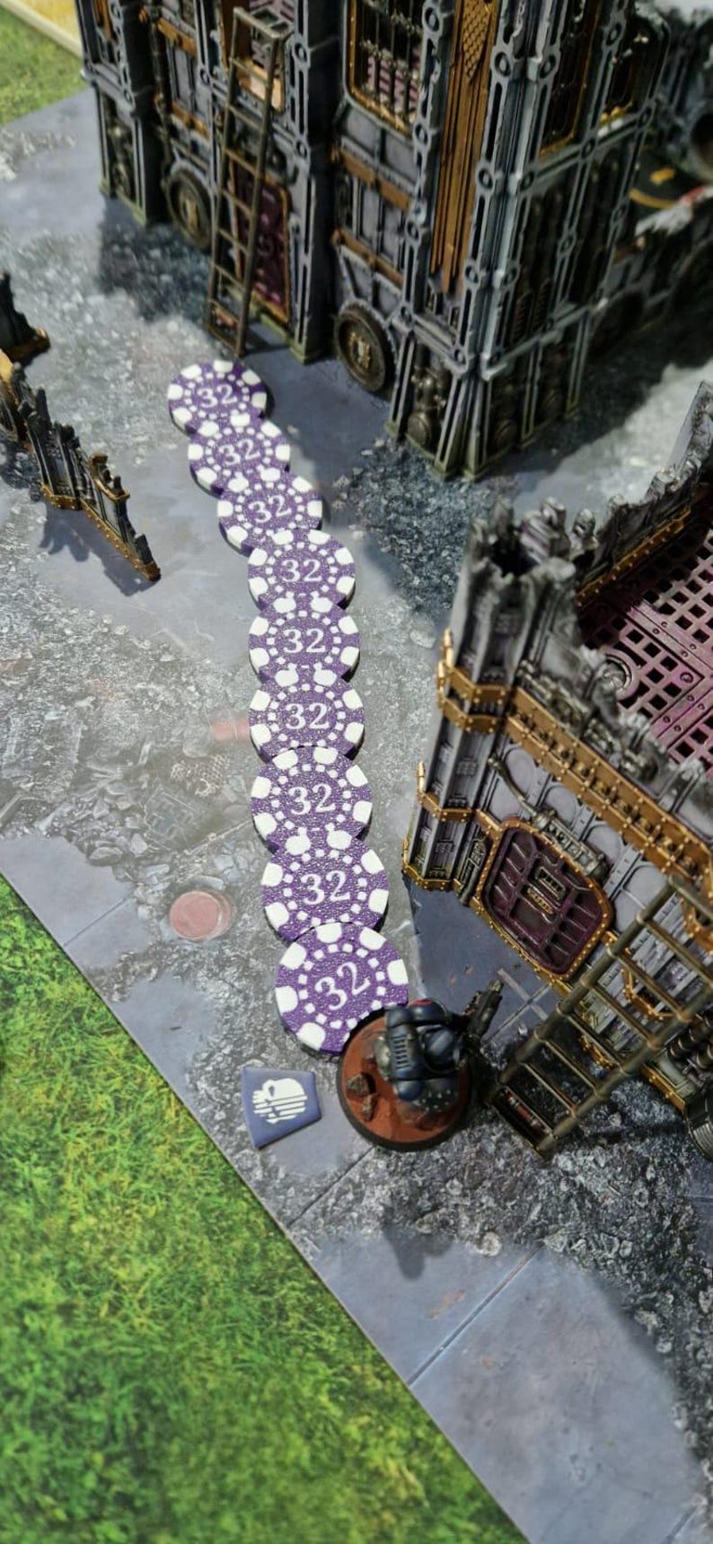 May include: A line of purple and white poker chips with the number 32 printed on each chip. The chips are arranged in a line on a gray surface. A miniature figure of a soldier is standing on a brown base next to the chips.
