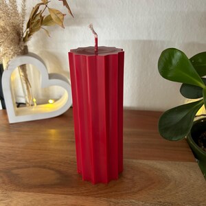 May include: A tall, red pillar candle with a geometric, ridged design. The candle has a wick at the top and stands on a wooden surface. Decorative elements, including a heart-shaped object and greenery, are in the background.