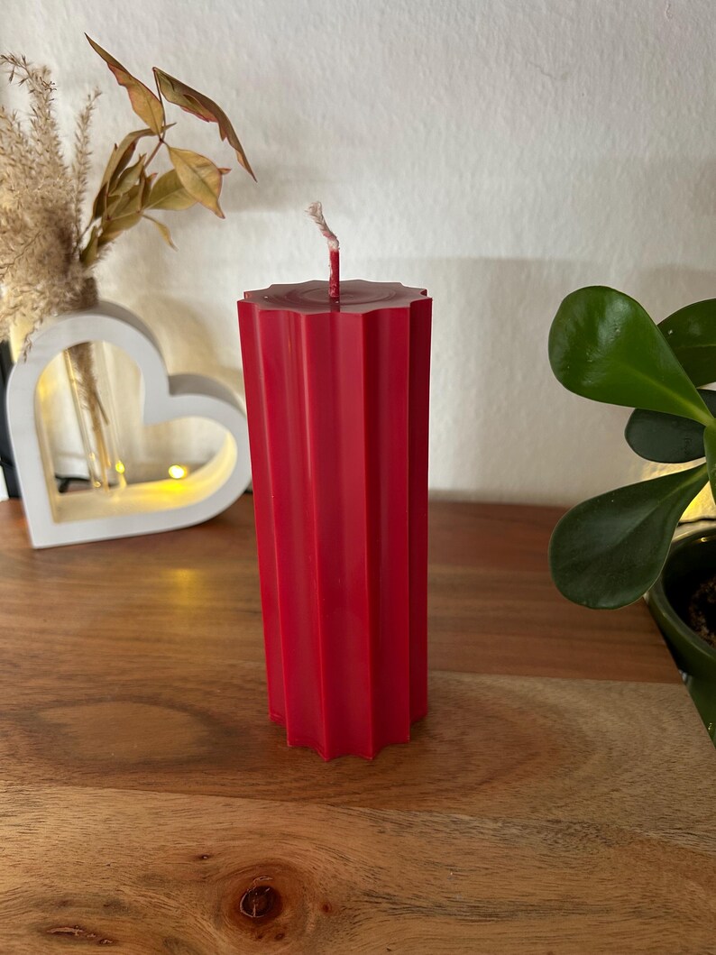 May include: A tall, red pillar candle with a geometric, ridged design. The candle has a wick at the top and stands on a wooden surface. Decorative elements, including a heart-shaped object and greenery, are in the background.