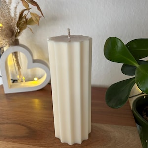 May include: Ivory-colored pillar candle with a fluted design. The candle has a geometric top and a wick. It is placed on a wooden surface, with a decorative heart-shaped light and greenery in the background.