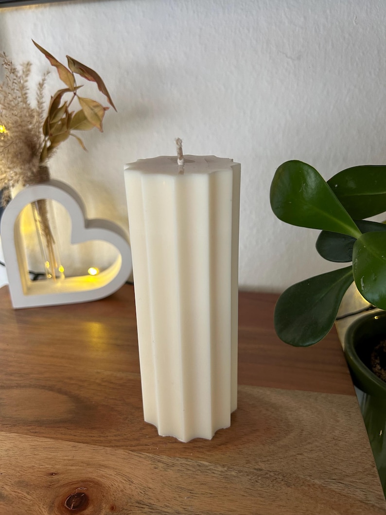May include: Ivory-colored pillar candle with a fluted design. The candle has a geometric top and a wick. It is placed on a wooden surface, with a decorative heart-shaped light and greenery in the background.
