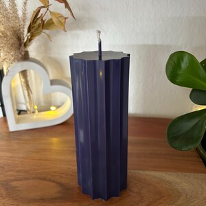 May include: A dark blue, fluted pillar candle with a wick. The candle has a starburst-shaped top and is displayed on a wooden surface. Decorative elements, including a heart-shaped light and plants, are in the background.