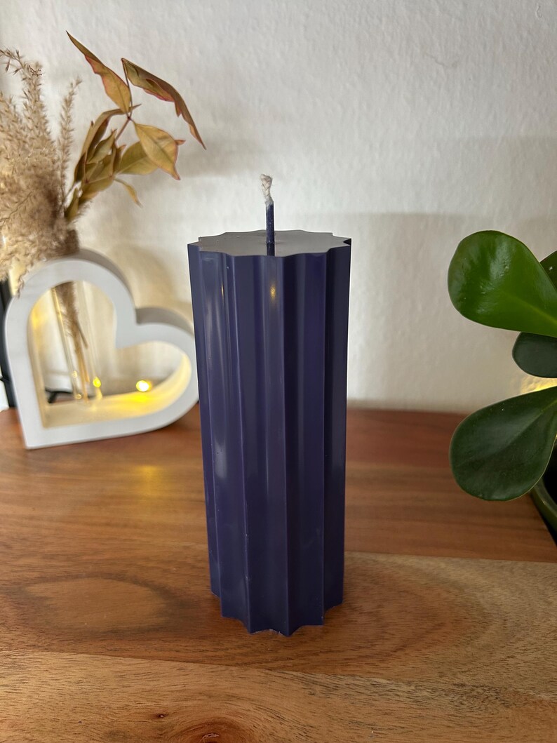 May include: A dark blue, fluted pillar candle with a wick. The candle has a starburst-shaped top and is displayed on a wooden surface. Decorative elements, including a heart-shaped light and plants, are in the background.