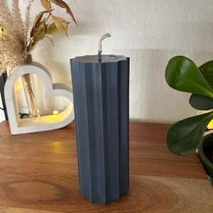 May include: A dark teal pillar candle with a textured, fluted design. The candle has a square top and a wick. The candle is on a wooden surface. Decorative home decor item.