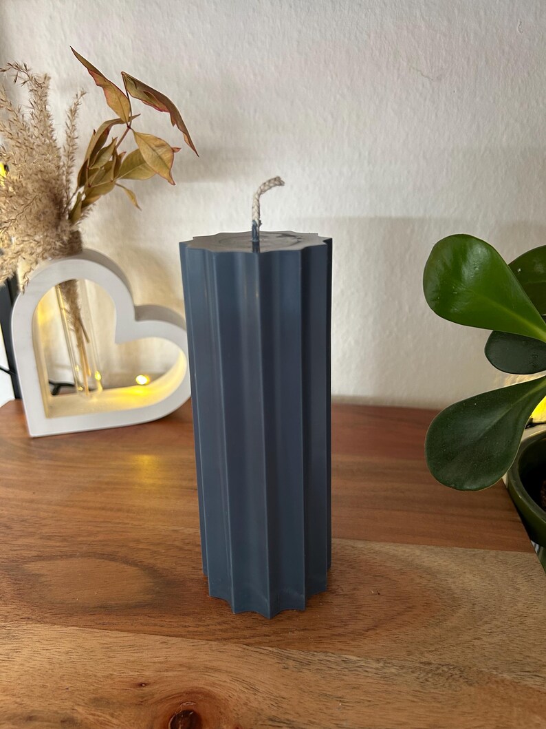 May include: A dark teal pillar candle with a textured, fluted design. The candle has a square top and a wick. The candle is on a wooden surface. Decorative home decor item.