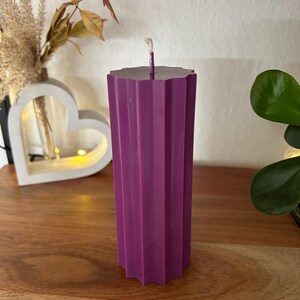 May include: A tall, purple pillar candle with a geometric, ridged design. The candle has a flat, star-shaped top with a white wick. The candle is displayed on a wooden surface, with decorative elements in the background.