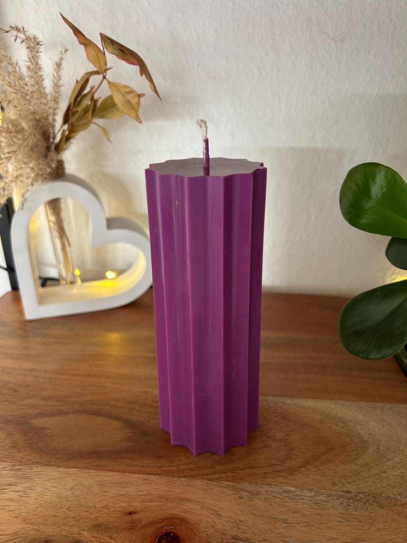 May include: A tall, purple pillar candle with a geometric, ridged design. The candle has a flat, star-shaped top with a white wick. The candle is displayed on a wooden surface, with decorative elements in the background.