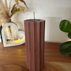 May include: A tall, brown pillar candle with a textured, fluted design. The candle has a star-shaped top and a wick. The candle is on a wooden surface, with decorative plants in the background.