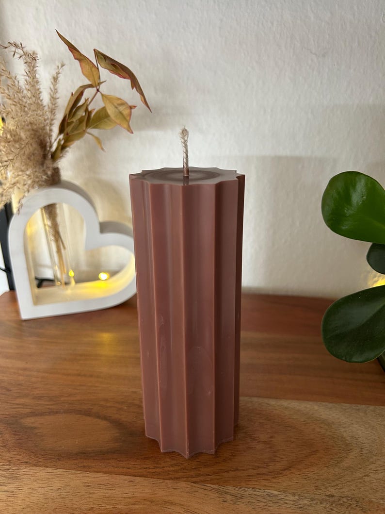 May include: A tall, brown pillar candle with a textured, fluted design. The candle has a star-shaped top and a wick. The candle is on a wooden surface, with decorative plants in the background.