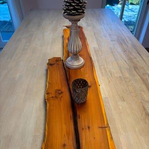 May include: A rustic wooden slab with a natural edge, featuring a light brown and orange colour. A decorative white ceramic pedestal with a pine cone sits on the slab, along with a metal candle holder.
