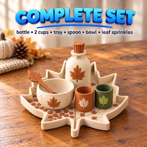 Puede incluir: Un conjunto completo que incluye una bandeja con forma de hoja, una botella con tapón, dos tazas, un cuenco, una cuchara y adornos de hojas. Los artículos son de color crema, naranja y verde con motivos de hojas. El texto COMPLETE SET bottle 2 cups tray spoon bowl leaf sprinkles es visible.