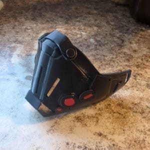 Puede incluir: Una máscara negra y angular con detalles circulares rojos y detalles dorados. La máscara tiene un diseño curvo, con una correa y varias aberturas. La máscara parece estar hecha de un material duro, posiblemente plástico.