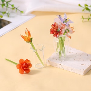 May include: A still life arrangement featuring miniature glass flowers in a clear vase and a small glass bud vase. The flowers are orange, red, pink, and purple. One flower is lying on the surface. The arrangement is on a white textured block.