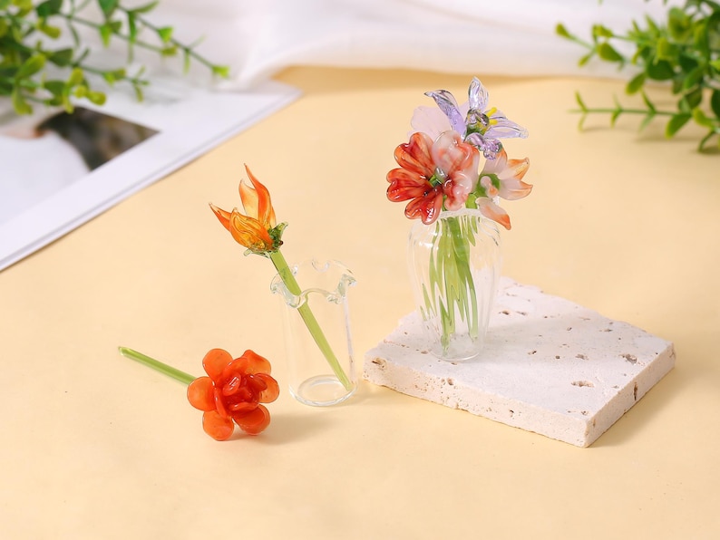 May include: A still life arrangement featuring miniature glass flowers in a clear vase and a small glass bud vase. The flowers are orange, red, pink, and purple. One flower is lying on the surface. The arrangement is on a white textured block.