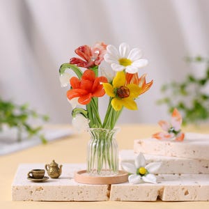 May include: A glass flower bouquet in a clear vase. The bouquet includes orange, yellow, and white flowers with green stems. A miniature tea set and a single flower are also present.