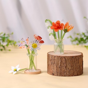 May include: Two glass flower arrangements. One with orange and white flowers in a clear vase on a wood slice. The other has a daisy and other flowers in a small vase on a wooden base. Decorative floral art.