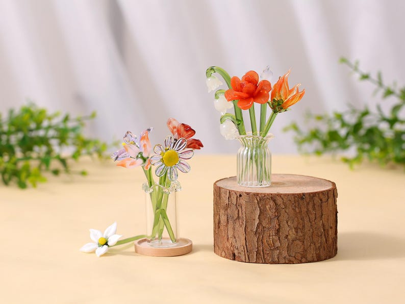 May include: Two glass flower arrangements. One with orange and white flowers in a clear vase on a wood slice. The other has a daisy and other flowers in a small vase on a wooden base. Decorative floral art.