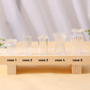 May include: Five clear glass vases of different shapes and sizes are displayed on a small wooden shelf. Each vase is labelled with a number. The vases have different designs, including ribbed and fluted patterns, and a ruffled top.