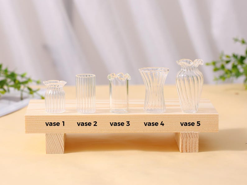 May include: Five clear glass vases of different shapes and sizes are displayed on a small wooden shelf. Each vase is labelled with a number. The vases have different designs, including ribbed and fluted patterns, and a ruffled top.