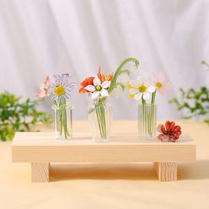 May include: Three small glass vases with colourful artificial flowers sit on a light wood platform. The flowers include daisies, poppies, and other blooms. A small, red flower is placed on the platform.