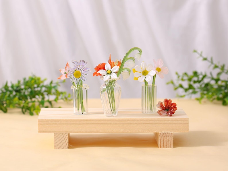 May include: Three small glass vases with colourful artificial flowers sit on a light wood platform. The flowers include daisies, poppies, and other blooms. A small, red flower is placed on the platform.