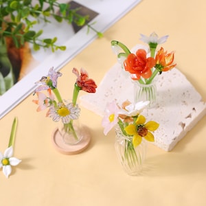 May include: Miniature glass flower bouquets in clear vases. The bouquets feature colourful flowers in shades of red, orange, yellow, pink, and white. One flower is separate. The vases sit on a beige surface.